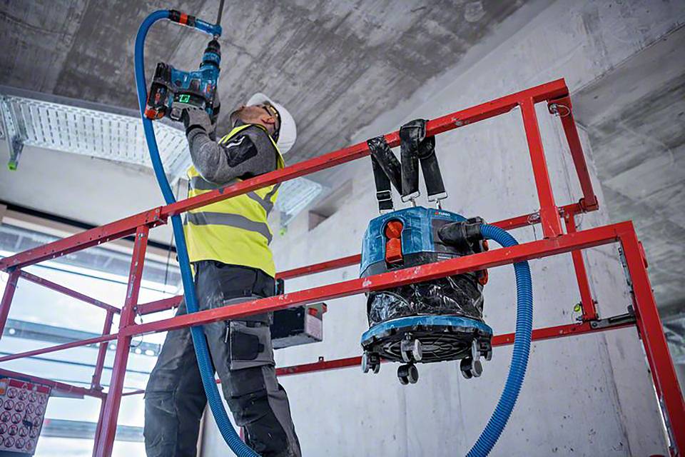 Arbeiter auf Baugerüst verwendet Bohrmaschine, trägt Schutzkleidung und -helm. Staubsauger an Gerüst befestigt, moderner Bauprozess.