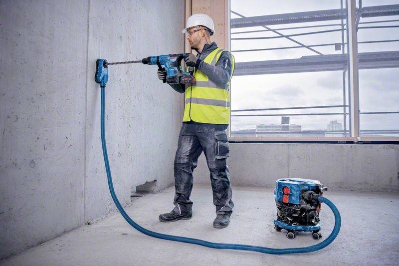 Eine Person in Arbeitskleidung bohrt in eine Betonwand auf einer Baustelle. Ein Staubsauger ist an die Bohrmaschine angeschlossen.