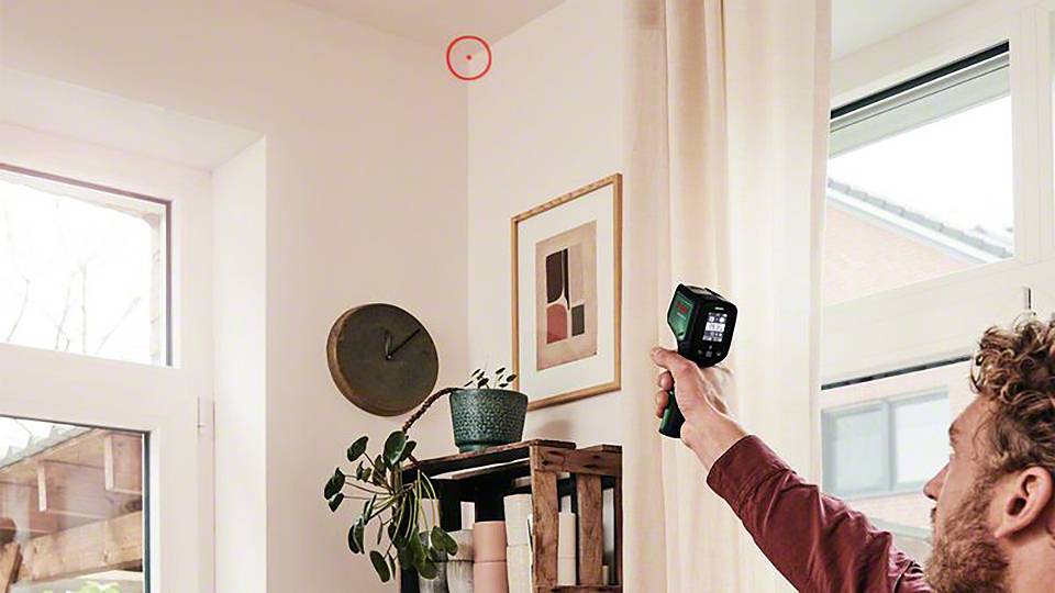 Eine Person scannt eine Wandecke mit einem Wärmebildgerät in einem Wohnraum. Ein roter Kreis markiert eine Stelle an der Decke.