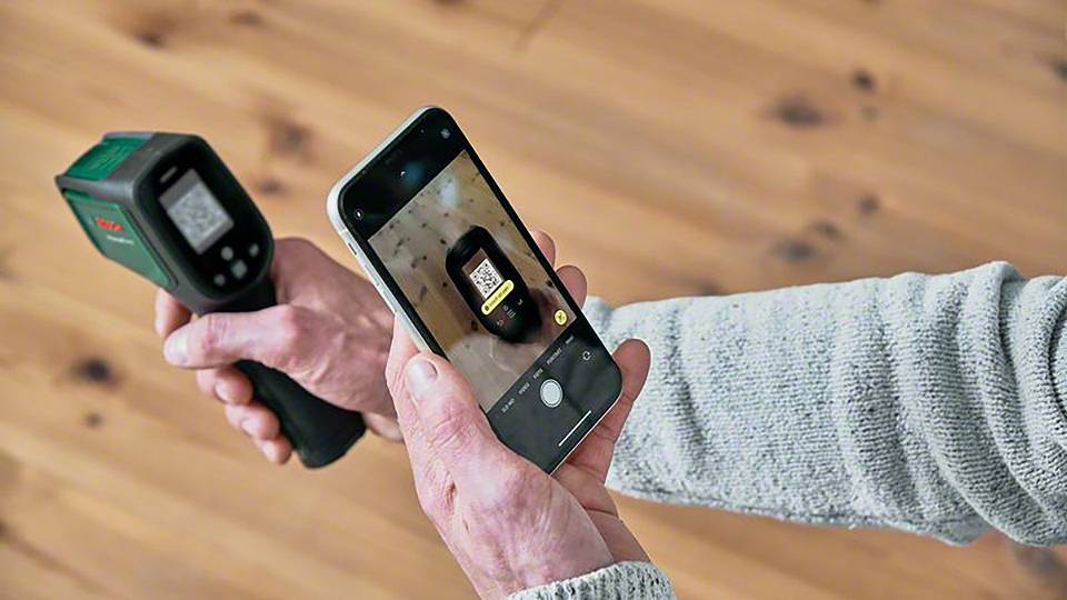 Eine Person hält ein Infrarot-Thermometer und ein Smartphone, das den Bildschirm des Thermometers fotografiert. Hintergrund ist Holzboden.