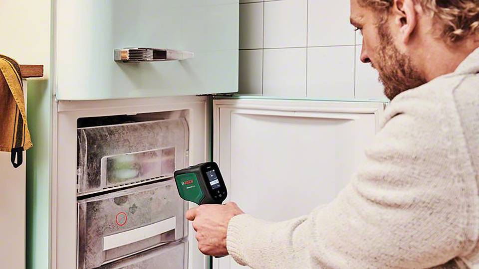 Ein Mann benutzt ein Infrarotthermometer, um die Temperatur des Gefrierschranks zu messen. Der Gefrierschrank ist geöffnet und vereist.
