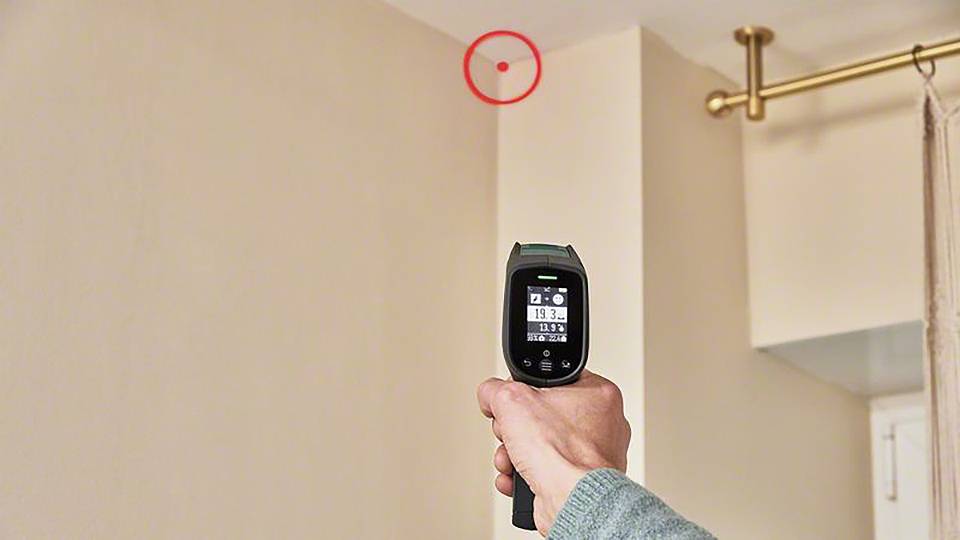 Eine Person zielt mit einem Infrarot-Thermometer auf eine Zimmerdecke. Ein roter Kreis markiert einen Punkt an der Decke.