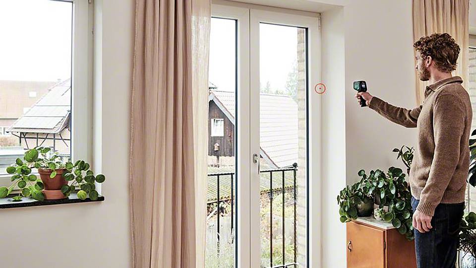 Ein Mann misst mit einem Gerät die Isolierung an einem großen Fenster. Pflanzen stehen auf der Fensterbank und einem Schrank.