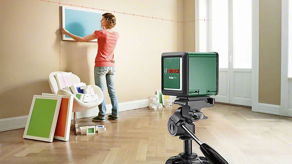 Eine Person verwendet ein Laser-Nivelliergerät, um Rahmen an einer Wand auszurichten. Auf dem Boden liegen mehrere Farbproben und Bilder.