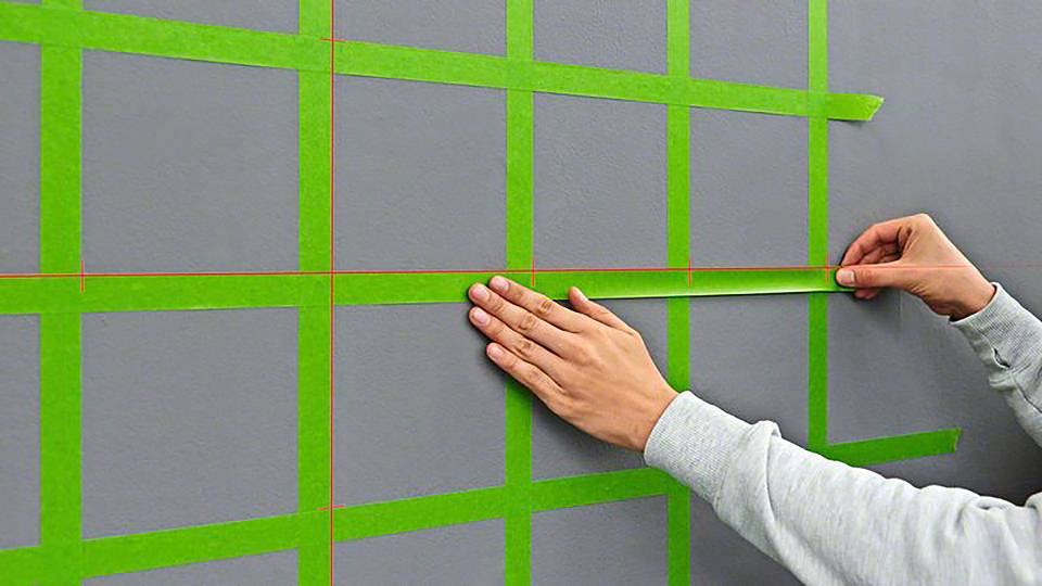 Eine Person klebt grünes Malerkreppband in einem Gittermuster auf eine graue Wand.