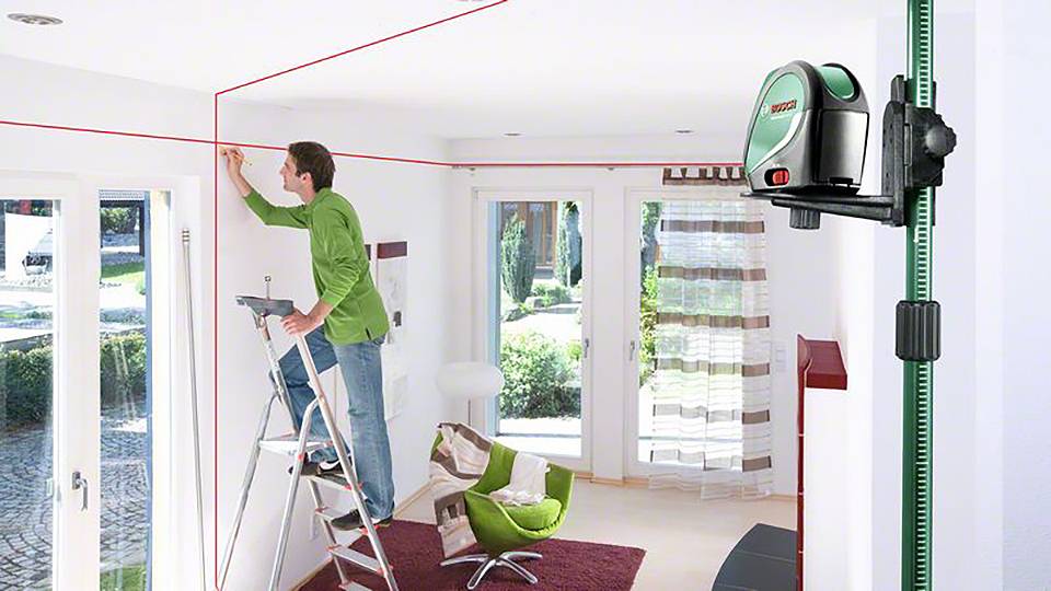 Ein Mann misst mit einem Laser-Nivelliergerät und roten Linien an der Decke, während er auf einer Leiter in einem hellen Wohnzimmer arbeitet.