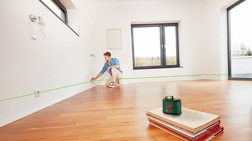 Eine Person im blauen Hemd justiert einen Laser an der Wand eines leeren, hellen Raumes mit Holzfußboden. Ein Lasergerät steht auf Büchern.