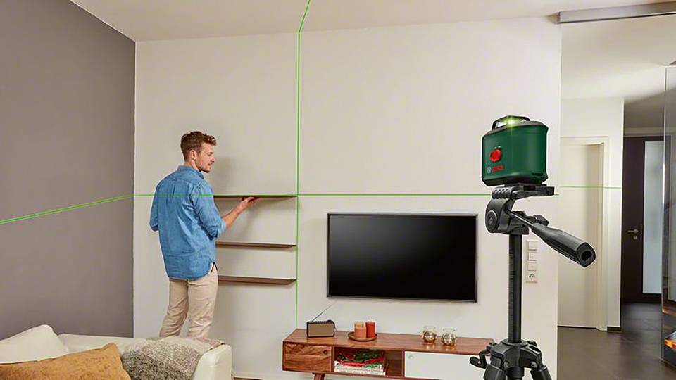Ein Mann richtet ein Regal mit einem grünen Laser-Nivelliergerät im Wohnzimmer aus. Im Hintergrund ein Fernseher und ein Sofa.