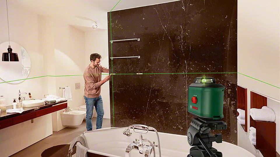 Ein Mann verwendet einen grünen Laser-Multilinienlaser in einem Badezimmer, um Fliesen an einer braunen Wand präzise auszurichten.