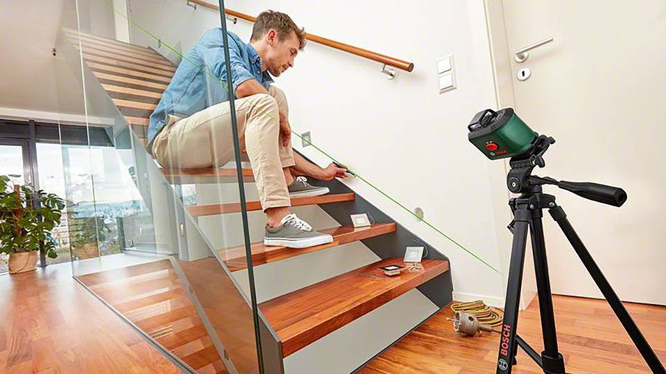 Ein Mann misst mit einem Laser auf einer Holztreppe. Ein Stativ mit Gerät steht daneben. Moderne, helle Umgebung.