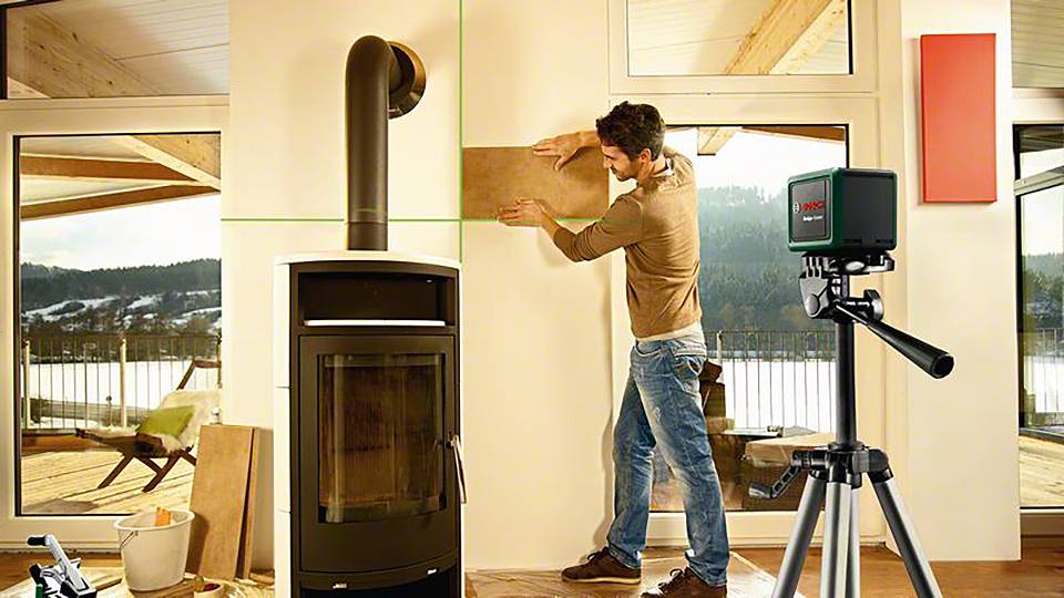 Ein Mann misst eine Wand neben einem Holzofen mit einem Lasergerät. Draußen ist eine Winterlandschaft zu sehen.