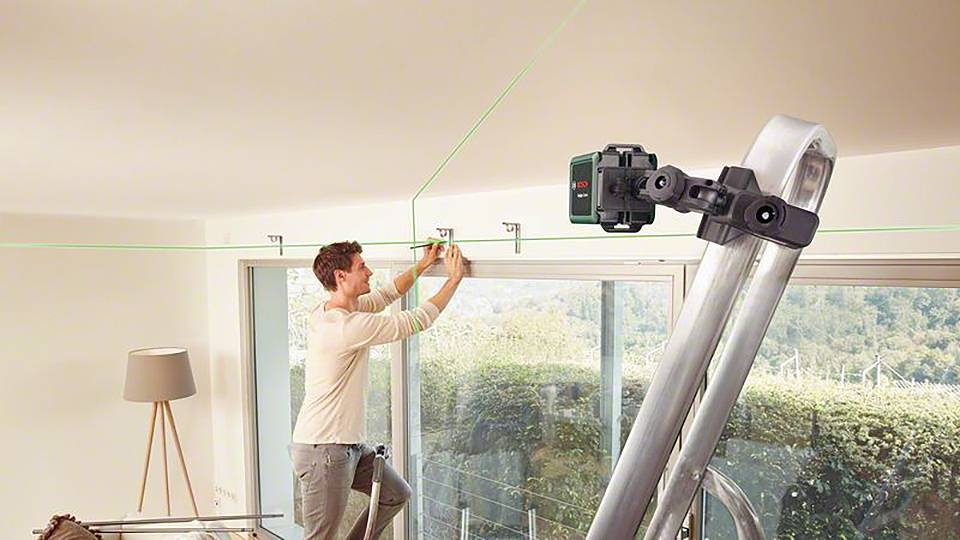 Ein Mann montiert Gardinenstangen an der Decke mit Hilfe eines Laserwerkzeugs zur präzisen Ausrichtung. Eine Leiter steht ihm zur Verfügung.