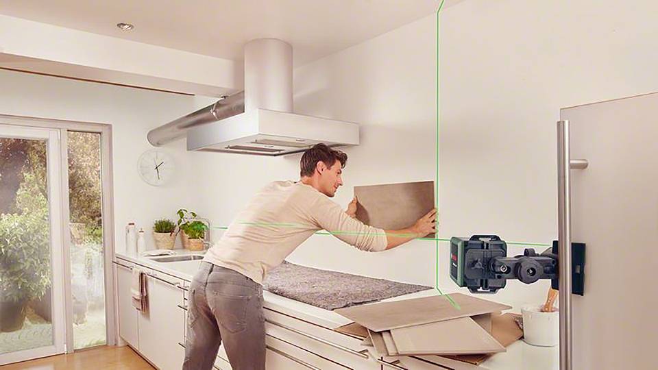 Ein Mann richtet eine Kachel an der Küchenwand aus, mit Hilfe eines grünen Laser-Nivelliergeräts. Die Küche ist modern eingerichtet.