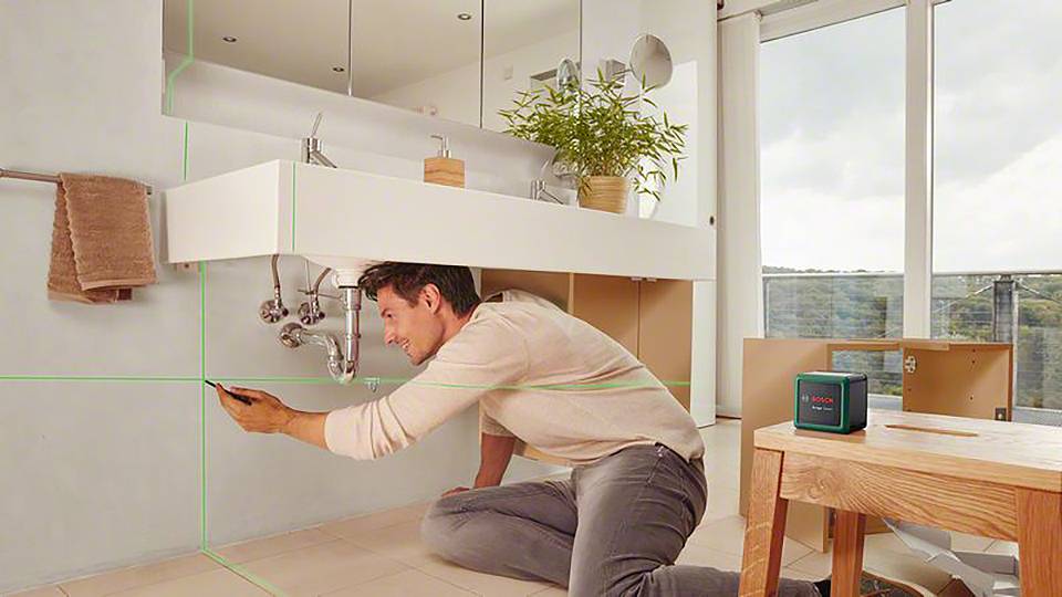 Ein Mann justiert einen Laserentfernungsmesser im Badezimmer, während er sich vor einem Waschbecken befindet. Ein grüner Laser projiziert Linien an die Wand.