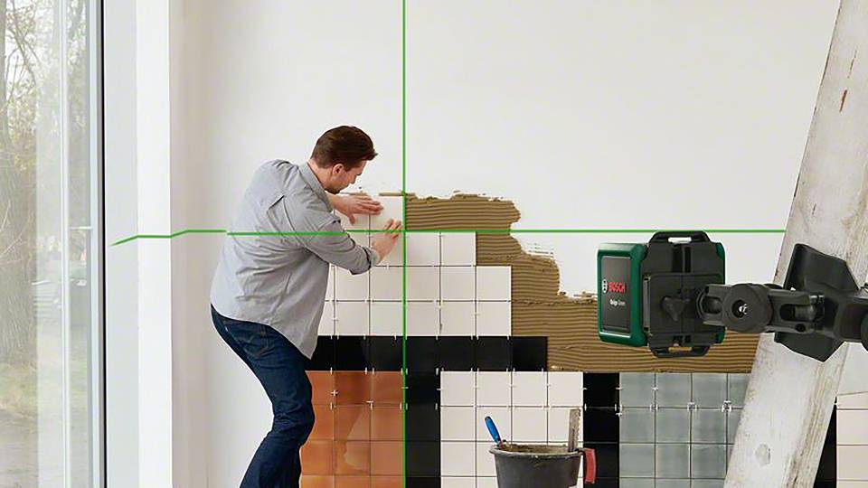 Ein Mann klebt Fliesen an eine Wand mit einem Lasernivelliergerät daneben. Eine Leiter steht rechts; ein Eimer Mörtel ist darunter.