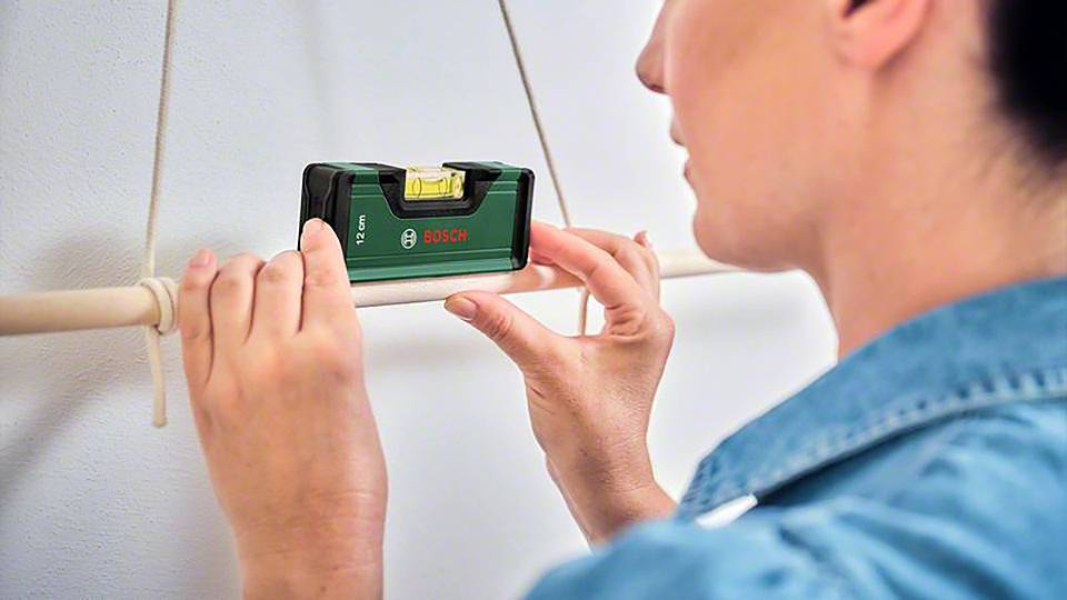 Eine Person richtet eine Wasserwaage an einer Holzstange aus, um sie gerade zu hängen. Die Wasserwaage zeigt das Bosch-Logo.