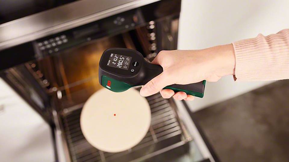 Eine Person hält ein Infrarot-Thermometer über eine geöffnete Ofentür, um die Temperatur eines Pizzasteins zu messen.