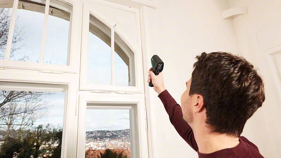 Ein Mann misst die Temperatur nahe eines Fensters mit einem Infrarotthermometer. Links ist ein Blick nach draußen auf bewaldete Hügel.