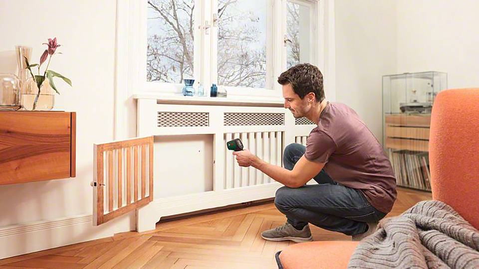 Ein Mann misst die Temperatur an einem Heizkörper in einem modernen Wohnzimmer. Im Hintergrund sind Fenster mit Blick auf Bäume.