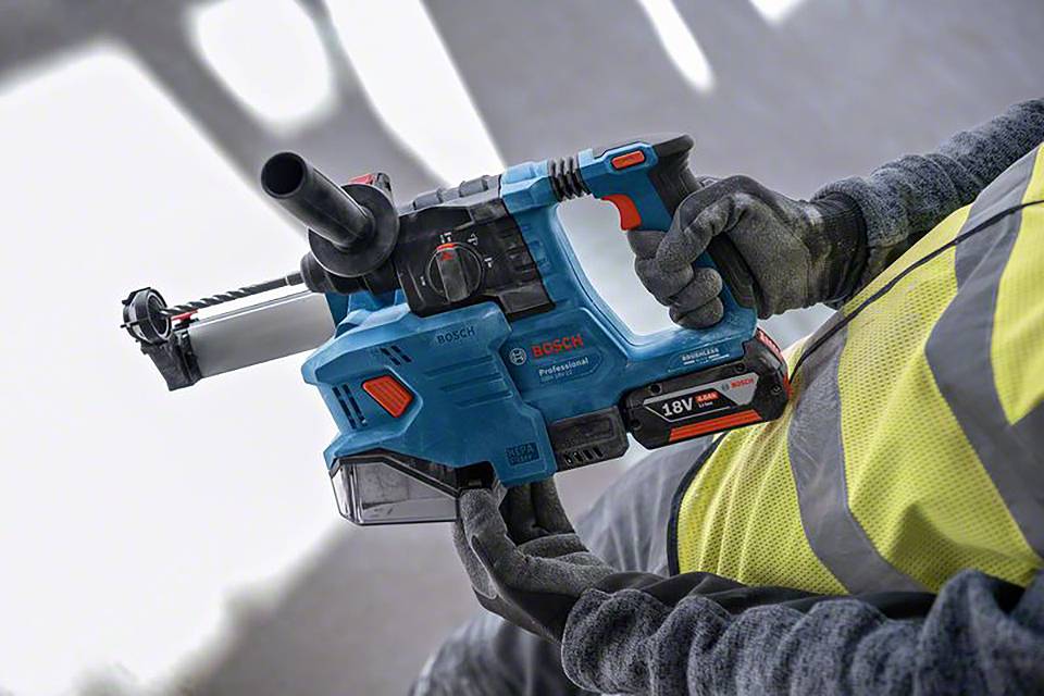 Person hält eine blaue Bosch-Bohrmaschine in der rechten Hand, trägt eine gelbe Sicherheitsweste und Arbeitshandschuhe.