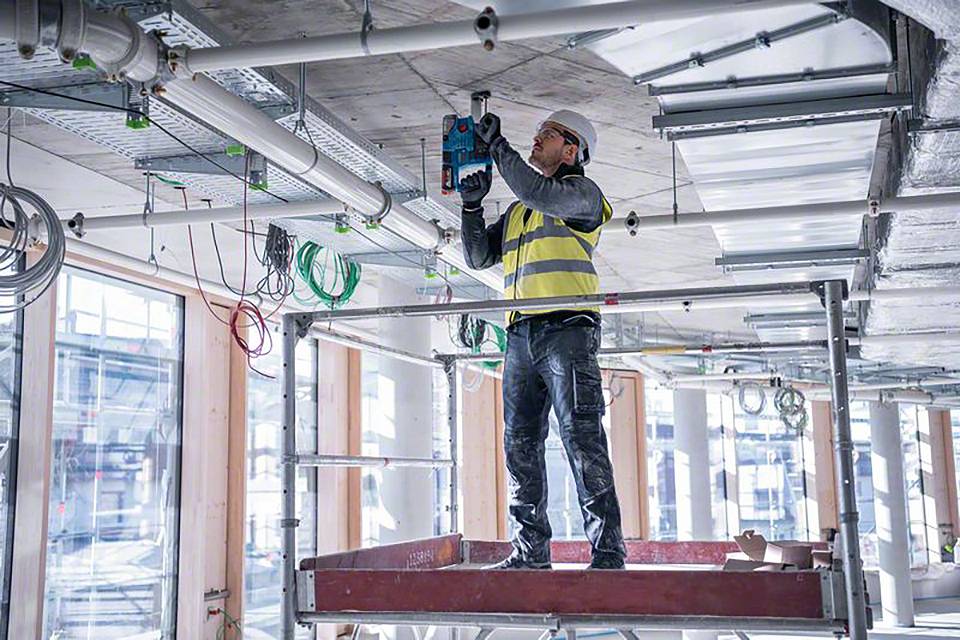 Arbeiter in Warnweste und Helm installiert Elektrik an der Decke einer Baustelle, umgeben von Baugerüsten und Rohren.