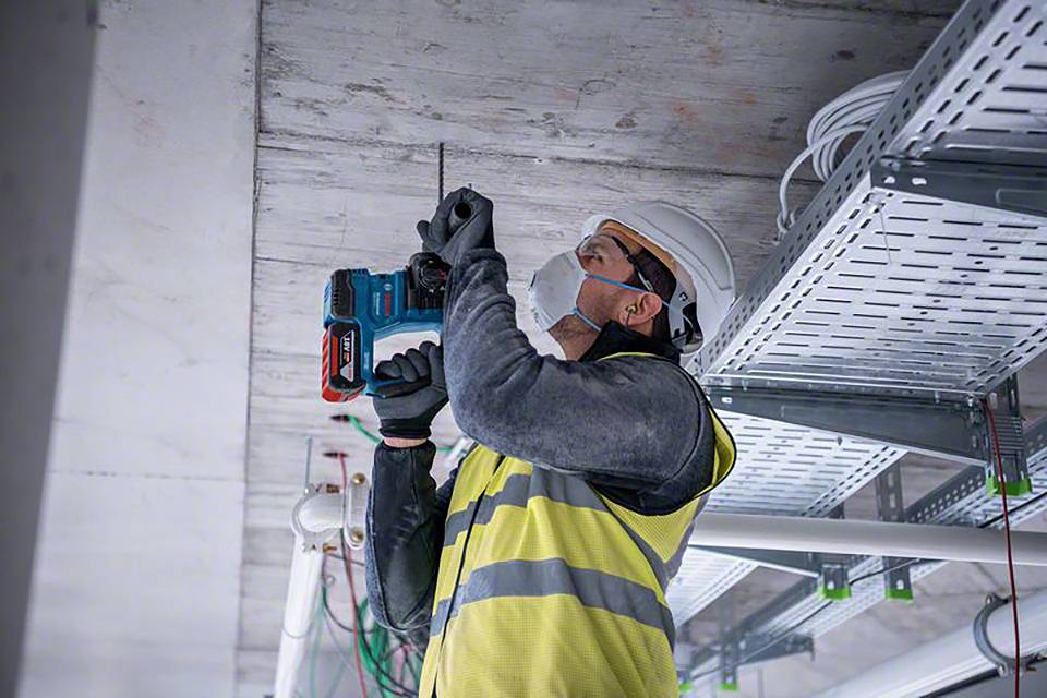 Ein Bauarbeiter mit Schutzhelm und Weste bohrt in die Decke eines Gebäudes. Im Hintergrund sind Kabelträger und Rohre sichtbar.
