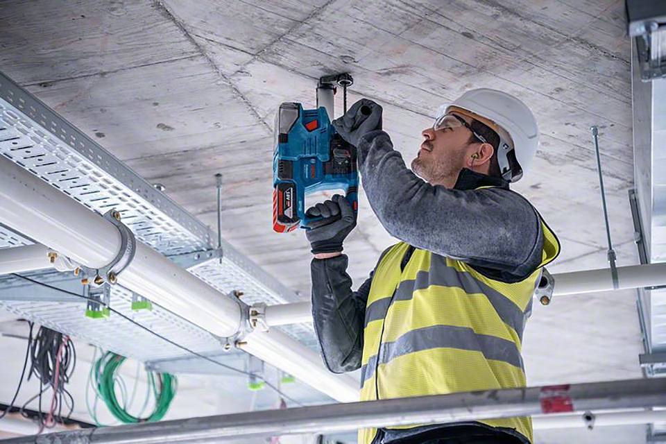 Ein Bauarbeiter in Schutzkleidung befestigt mit einer Bohrmaschine Kabel an der Decke einer Baustelle.