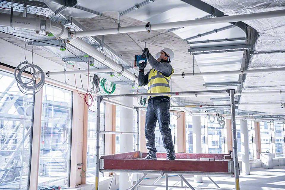 Ein Bauarbeiter in Schutzkleidung steht auf einem Gerüst und führt Installationsarbeiten an der Decke in einem unfertigen Gebäude durch.