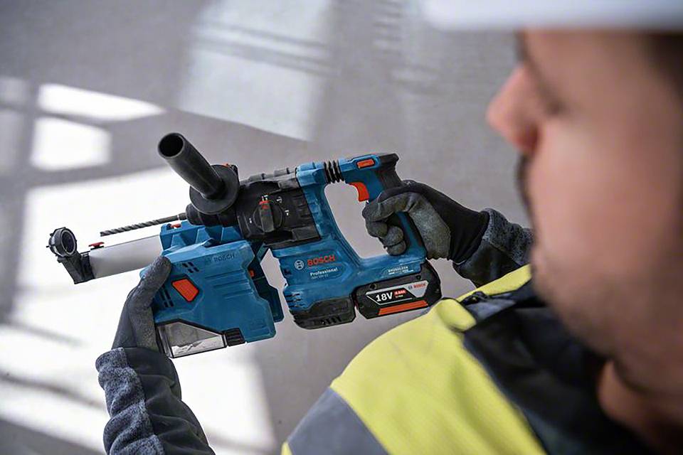 Eine Person hält einen blauen Bohrhammer. Der Hintergrund ist unscharf und die Person trägt eine Sicherheitsweste und Helm.