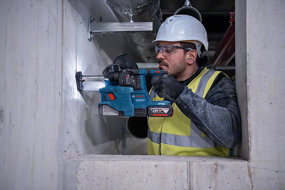 Ein Bauarbeiter trägt Helm und Schutzbrille, benutzt eine Bohrmaschine, um in eine Betonwand zu bohren, zeigt konzentrierte Arbeit.