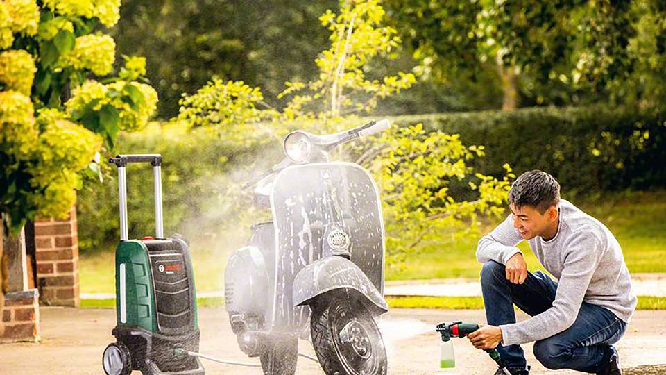 Ein Mann reinigt mit einem Hochdruckreiniger einen Roller im Freien. Im Hintergrund sind Bäume und Büsche zu sehen.