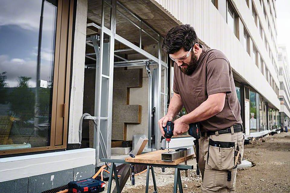 Ein Bauarbeiter bohrt mit einer Bohrmaschine ein Metallstück auf einer Baustelle neben einem Gebäude. Er trägt Schutzbrille und Werkzeuggürtel.