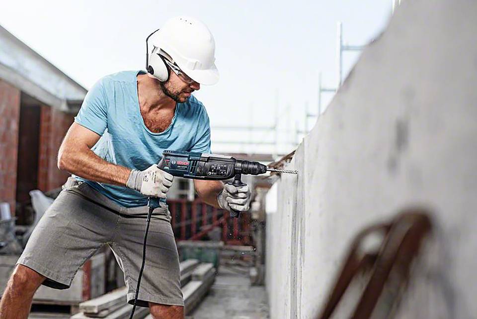 Ein Bauarbeiter trägt Schutzhelm und -brille und benutzt eine elektrische Bohrmaschine, um ein Loch in eine Betonwand zu bohren.