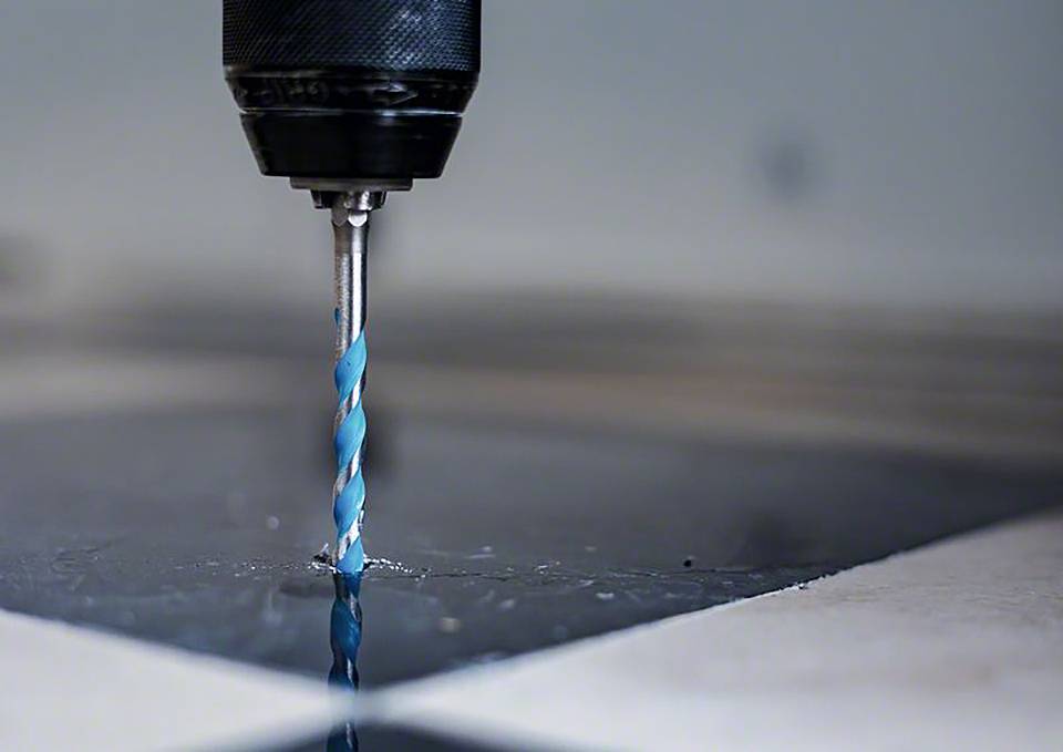 Ein Bohrer mit blau-weißer Spitze bohrt in eine schwarze Metalloberfläche. Der Bohrer dreht sich schnell und erzeugt Späne.