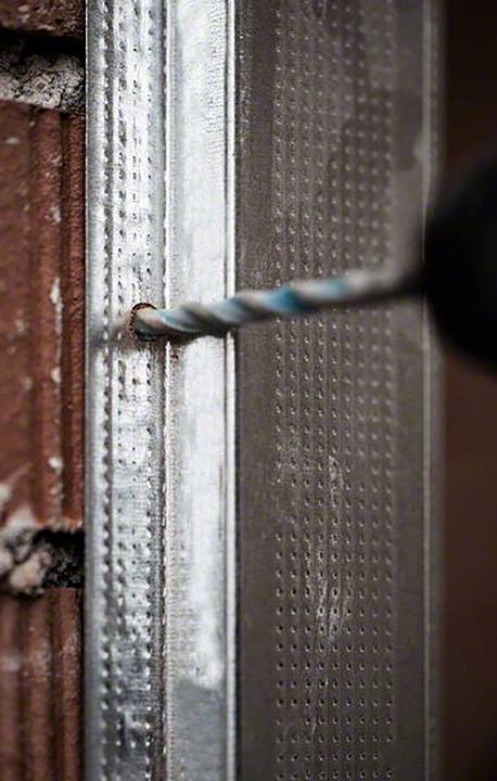 Ein Bohrer durchdringt ein Metallprofil an einer Backsteinwand. Fokus auf den Bohrer im Einsatz, zeigt Bau- oder Renovierungsarbeiten.