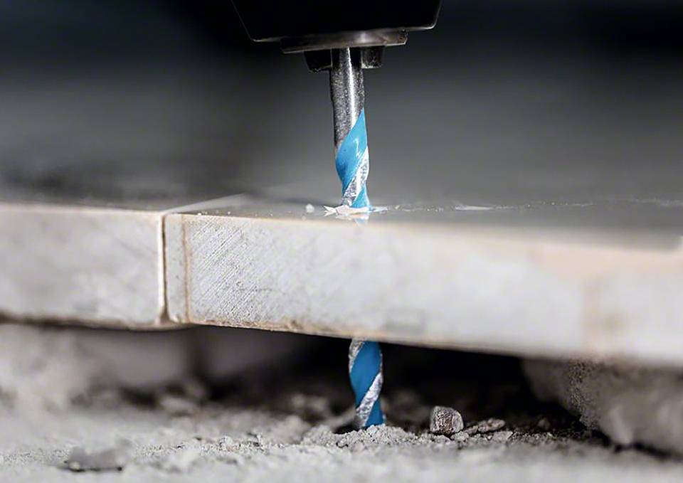 Ein blaues Bohrwerkzeug bohrt durch eine helle Fliese, umgeben von Staub und kleinen Trümmern auf einer Baustelle.