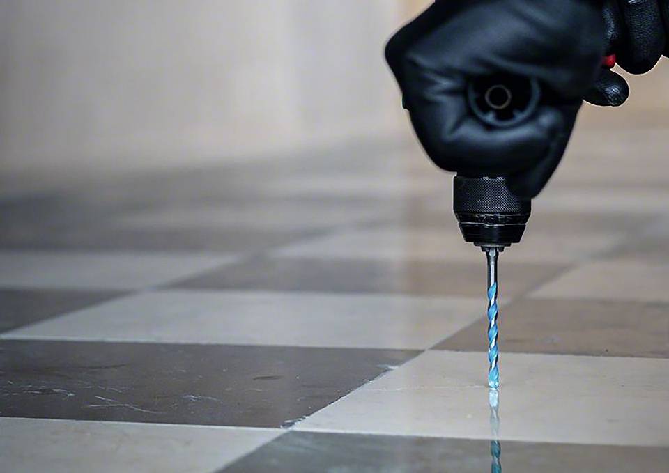 Eine Person in schwarzen Handschuhen hält einen Bohrer, der auf einen gefliesten Boden gerichtet ist. Das Bohrbit ist blau-schwarz.