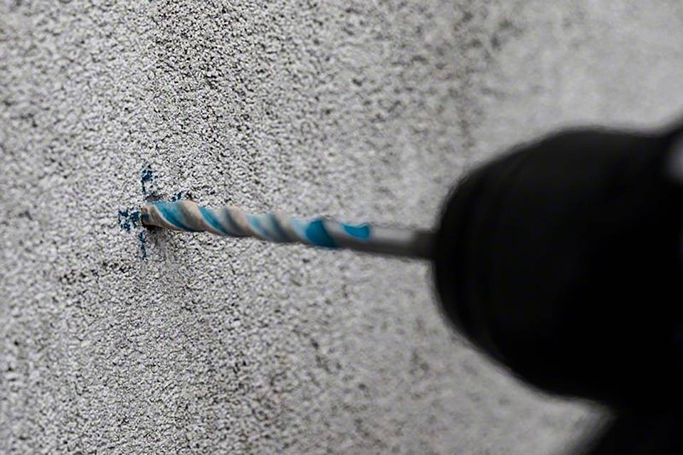 Ein elektrischer Bohrer bohrt in eine graue Betonwand. Der Bohrer dreht sich schnell und erzeugt Staub um das Bohrloch.