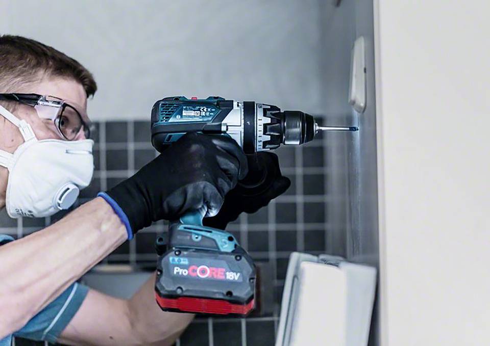 Ein Mann mit Schutzbrille und Maske benutzt eine elektrische Bohrmaschine, um ein Loch in eine geflieste Wand zu bohren.