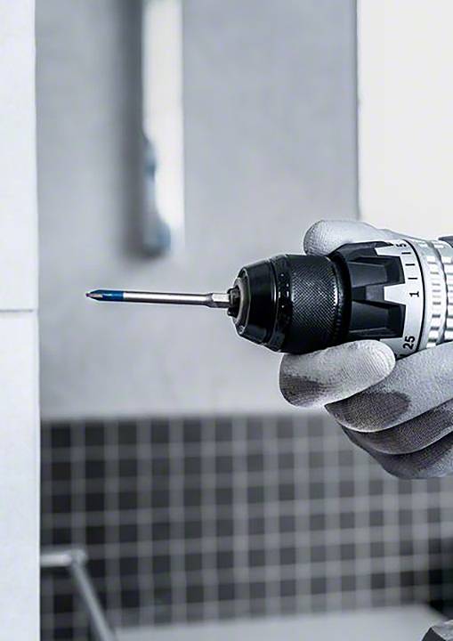 Eine Hand hält einen Bohrer, der bereit ist, um ein Loch in eine geflieste Wand zu bohren. Im Hintergrund ist unscharf ein Badezimmer erkennbar.