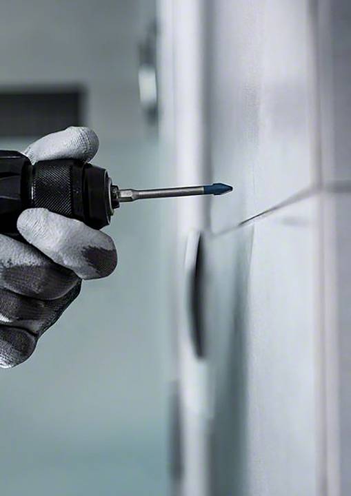 Eine Hand mit Handschuh hält einen Akkuschrauber, der gegen eine geflieste Wand gedrückt wird, um ein Loch zu bohren.