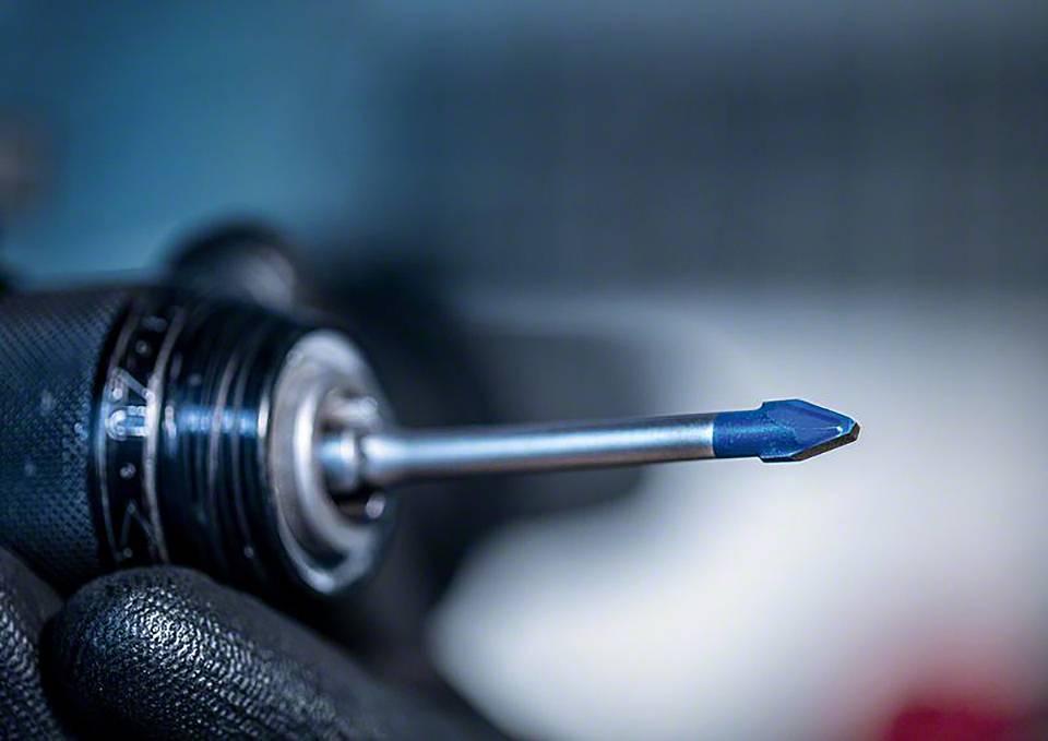 Ein Nahaufnahme einer Bohrmaschine mit blauem Bohrer, gehalten von einer Person mit schwarzen Arbeitshandschuhen.