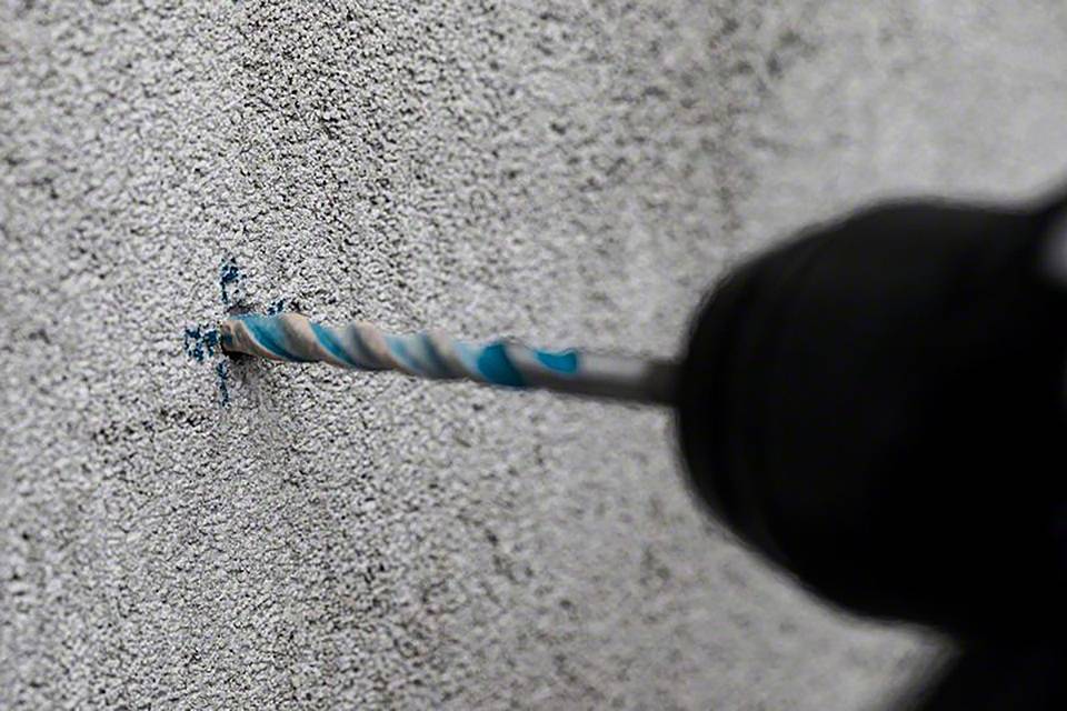 Eine Bohrmaschine mit blauem Bohrer durchbohrt eine graue, raue Betonwand. Staubpartikel fliegen um den Bohrer herum.