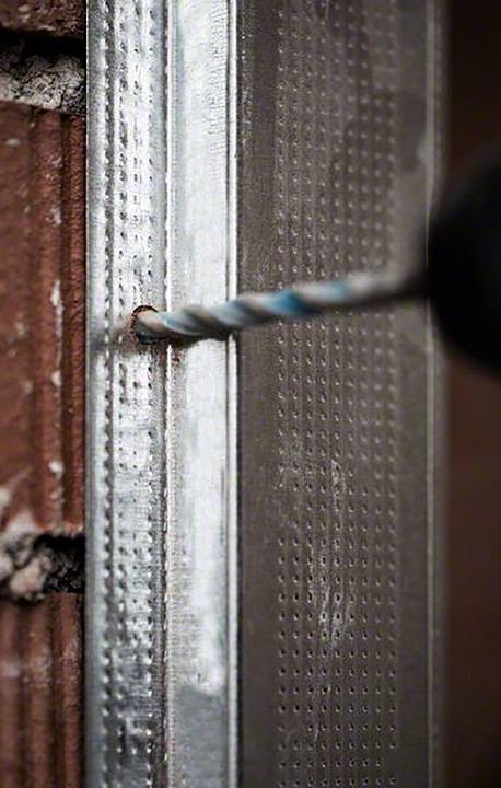 Ein Bohrer durchdringt ein Metallprofil an einer Backsteinwand.