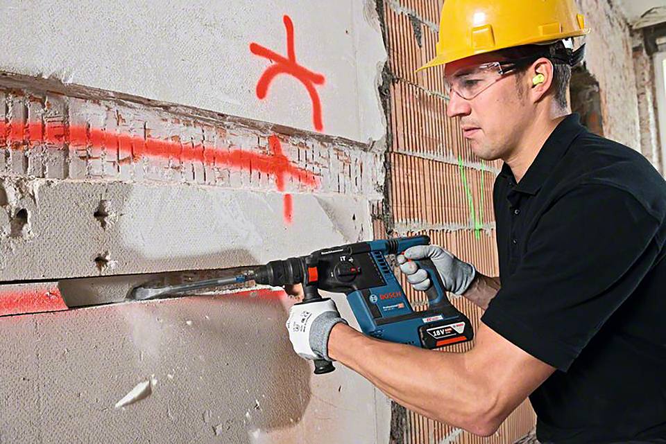 Ein Bauarbeiter in Schutzkleidung benutzt einen Bohrhammer, um eine Wand einzureißen. Rote Markierungen sind auf der Wand sichtbar.