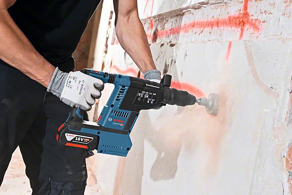 Ein Bauarbeiter benutzt einen elektrischen Bohrhammer, um Löcher in eine Betonwand zu bohren. Staub wirbelt um das Werkzeug herum auf.