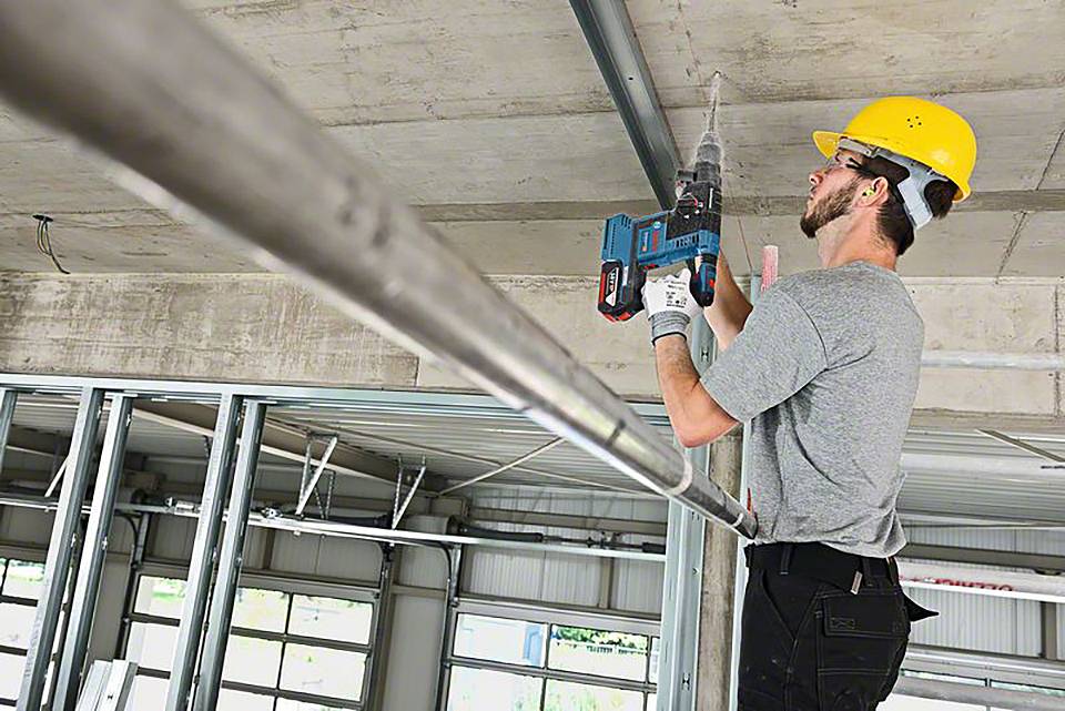 Ein Bauarbeiter in Schutzhelm und Gehörschutz verwendet eine Bohrmaschine, um an der Decke eines Gebäudes eine Metallleiste anzubringen.