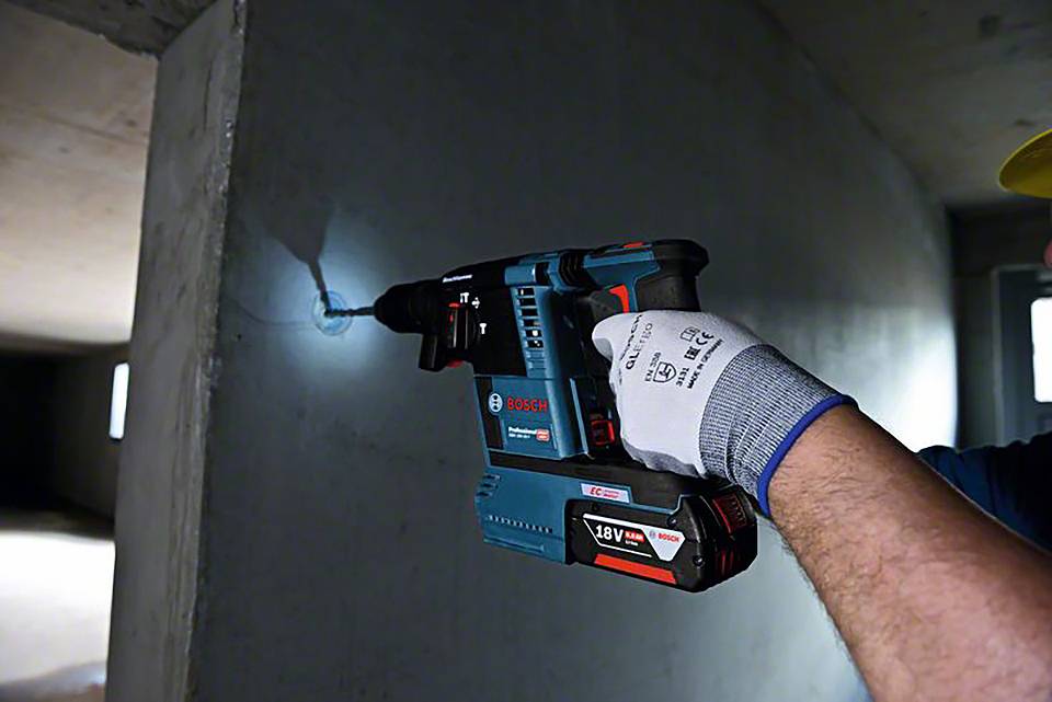 Eine Person verwendet einen blauen Bosch-Akkuschlagbohrhammer an einer Betonwand in einem Innenraum. Sie trägt einen Sicherheitshandschuh.
