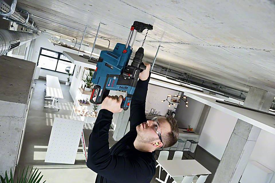 Eine Person in einer industriellen Umgebung bohrt mit einer großen, blauen Bohrmaschine in eine Betondecke. Im Hintergrund sind moderne Möbel.