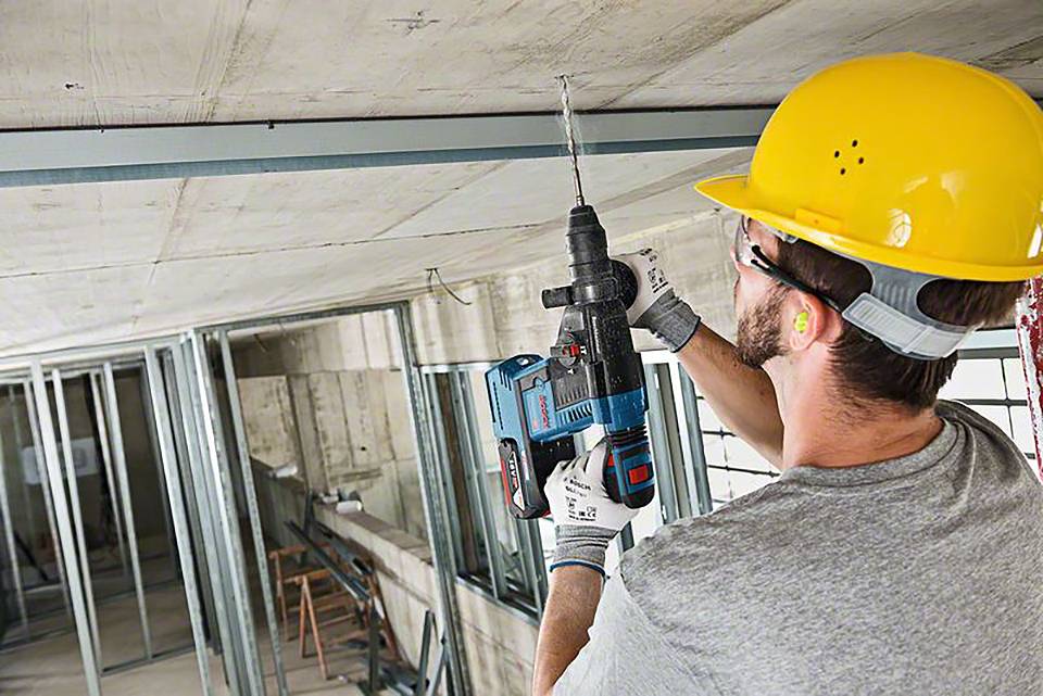 Ein Bauarbeiter mit Schutzhelm und Gehörschutz bohrt mit einer elektrischen Bohrmaschine in eine Decke.